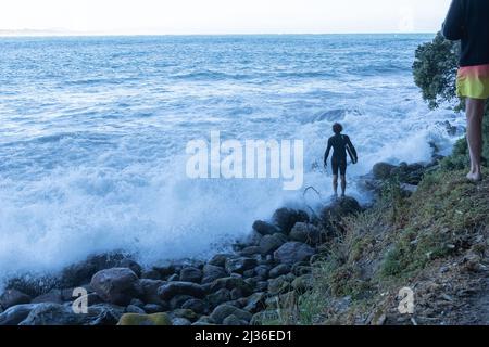 Tauranga Neuseeland - Januar 01 2022; Junge Rüde steht am felsigen Meeresrand und das Wellenbrett entscheidet, wann sie riskieren einzuspringen. Stockfoto