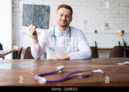 Porträt eines glücklichen männlichen Arztes in weißer Uniform, der Computertomographie des Gehirns untersucht und am Schreibtisch im Krankenhausbüro sitzt Stockfoto