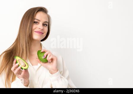 Porträt einer reizenden, lächelnden jungen, brünette kaukasischen Frau, die das weiße Hemd mit langen Haaren trägt und Avocado zeigt, isoliert auf weißem Hintergrund stehend Stockfoto