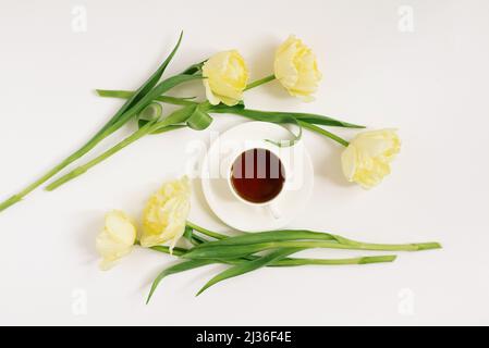 Eine weiße Porzellanschale mit schwarzem Tee auf einer Untertasse, umgeben von frühlingsgelben Tulpen auf weißem Hintergrund. Das Konzept des Frühlings, der Frauen und der Mutter Stockfoto