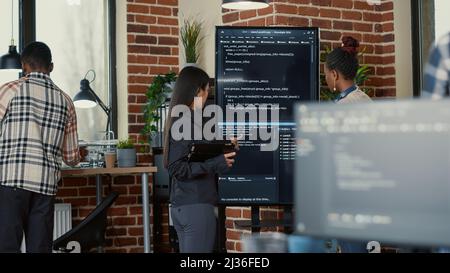 Softwareingenieur hält digitales Tablet, das Code auf dem Wandbildschirm analysiert, und erklärt dem Kollegen Programmer im IT-Startup-Büro Fehler. Team von Kodierern, die am Projekt der Innovationsgruppe ai zusammenarbeiten. Stockfoto
