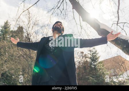 Attraktiver bärtiger lateinamerikanischer Mann, der mit ausgestreckten Armen glücklich in den Himmel blickt, im Outdoor-Medium-Shot-Wohlfühlkonzept. Hochwertige Fotos Stockfoto