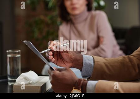 Nahaufnahme eines schwarzen Psychologen, der sich Notizen über die Patientin in der Zwischenablage machte, während sie ihr während der Therapie zuhörte Stockfoto