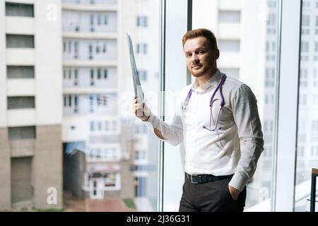 Porträt eines fokussierten männlichen Arztes in weißer Uniform, der Röntgenaufnahmen der Brust in der Nähe des Fensters im Krankenhausbüro untersucht Stockfoto