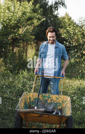 Lächelnder Bauer in Denim-Kleidung in der Nähe der Schubkarre mit Gartengeräten Stockfoto