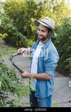 Glücklicher Gärtner im Hutkrempe, der Büsche mit einer Schere beschnitt Stockfoto