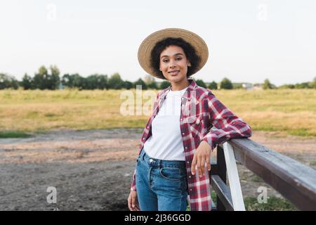 Hübsche afroamerikanische Frau im Strohhut lächelt vor der Kamera in der Nähe des Holzzauns auf dem Ackerland Stockfoto