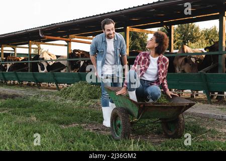 Fröhliche afroamerikanische Frau, die in Schubkarre sitzt und lächelnden Bauern anschaut Stockfoto