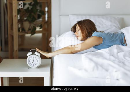 Frau schlafen auf dem Bett schaltet den Wecker am Morgen aufwachen, Selective Focus. Junge Frau greift, um Wecker auszuschalten, am frühen Morgen Stockfoto