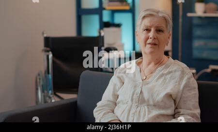 Porträt einer älteren Frau, die im Wohnzimmer auf die Kamera schaut. Ältere Menschen mit chronischer Behinderung genießen den Ruhestand und lächeln. Alter Erwachsener, der neben dem Rollstuhl sitzt, um den Transport zu machen. Stockfoto