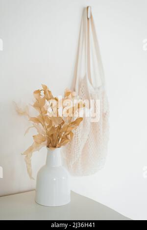 Eine getrocknete Blume in einer weißen Keramikvase und eine Stofftasche, die an der Wand im skandinavischen Dekor eines gemütlichen Zuhauses hängt Stockfoto