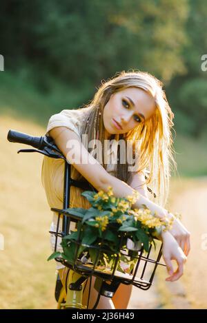 Eine junge, schöne Frau hält ein Fahrrad mit gelben Verbena-Blumen in einem Korb auf einem Spaziergang im Freien auf dem Land Stockfoto