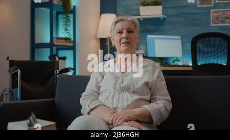 Porträt einer älteren Frau mit Körperverletzung, die auf der Couch sitzt. Person, die die Kamera anschaut und neben dem Rollstuhl im Wohnzimmer sitzt und Mobilitätshilfen für den Transport verwendet. Stockfoto