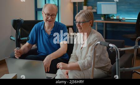 Ein Paar mit chronischer Behinderung, das nach dem Erfolg am Laptop fünf Punkte erzielt, Teamarbeit feiert und in den Ruhestand geht. Ältere Menschen mit Gehrahmen und Krücken, die am Computer Technik genießen. Stockfoto