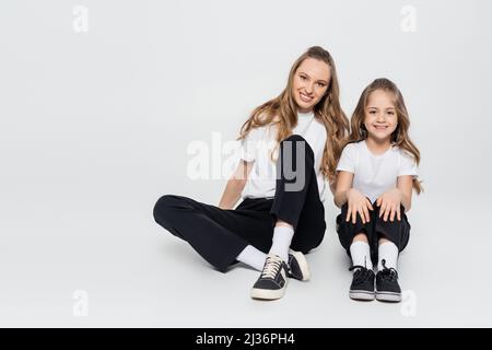 Fröhliche Frau und Mädchen in schwarz-weißen Kleidern sitzen auf grauem Hintergrund Stockfoto