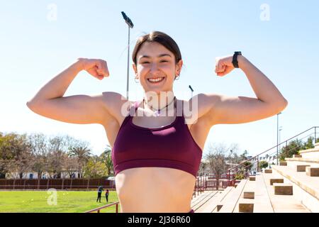 Eine junge kaukasische Sportlerin, die in einem Tank-Top gekleidet ist, übt Gewalt aus, wobei ihre Arme ihren Bizeps auf den Tribünen eines Sportbereichs zeigen. Stockfoto