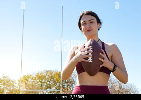 Eine junge kaukasische Athletin, die einen amerikanischen Fußballball an den Seilen hält. Sie trägt ein Aktivitätsarmband und in Th sind Torpfosten sichtbar Stockfoto