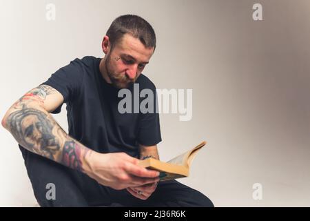 Hübscher bärtiger Kaukasischer tätowierte Mann, der auf dem Boden sitzt und die Heilige Bibel liest - Faith Konzept isoliert auf hellem Hintergrund mittlere Ganzaufnahme Studioaufnahme. Hochwertige Fotos Stockfoto