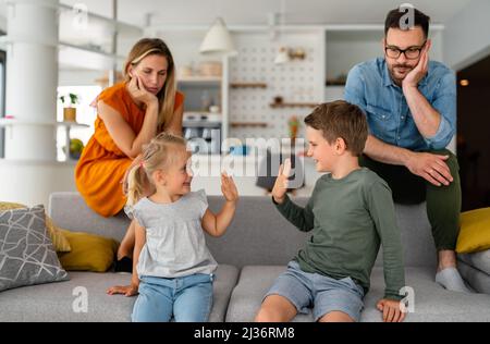 Müde Eltern, die auf der Couch sitzen, fühlen sich erschöpft genervt, während glückliche Kinder zusammen spielen. Stockfoto