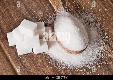 Granulatzucker in Holzlöffel und Zuckerwürfel in Pyramide auf Holzhintergrund gestapelt. Stockfoto