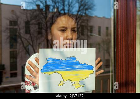 Ein Mädchen in einem gestickten Kleid blickt aus dem Fenster, hält ein weißes Blatt mit einem Bild und einer Inschrift. Das Mädchen protestiert gegen den Krieg.Ukraine Stockfoto