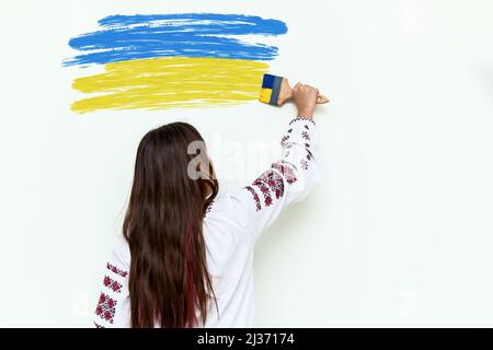 Ein Mädchen in einem Kleid mit Stickerei zeichnet an einer weißen Wand, hält einen Pinsel in blau und gelb gemalt. Das Mädchen protestiert gegen den Krieg und zieht die Flote Stockfoto