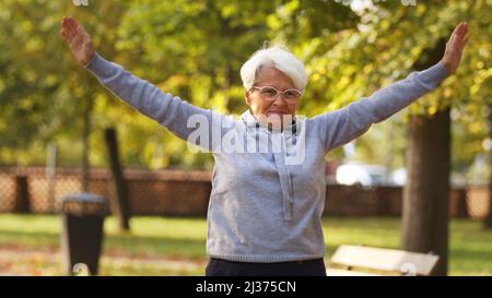 Ältere kaukasische Frau in einer Brille hält die Hände oben tun Sport Gymnastik im Park gesunde Lebensweise Konzept Mittelschuss selektive Fokus Park Hintergrund . Hochwertige Fotos Stockfoto