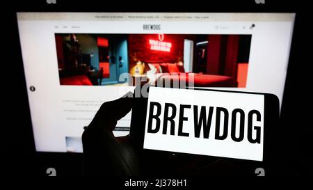 Person, die Mobiltelefon mit dem Logo des britischen Brauereiunternehmens BrewDog plc auf dem Bildschirm vor der Business-Webseite hält. Konzentrieren Sie sich auf die Telefonanzeige. Stockfoto