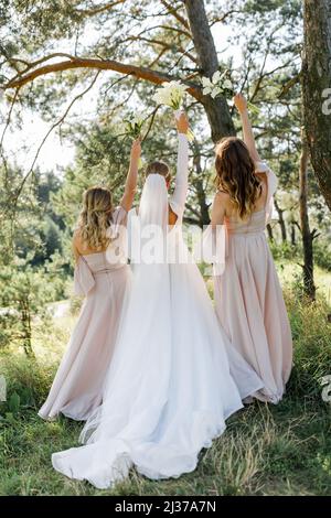 Zwei Brautjungfern in pudrigen Kleidern, die mit Blumensträußen in den Händen halten, stehen mit dem Rücken in einem weißen Kleid in der Nähe der Braut mit einem Brautstrauß in ihrem Rücken Stockfoto