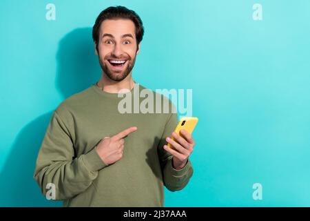 Foto von beeindruckt Brunet jungen Kerl zeigen Telefon tragen khaki Sweatshirt isoliert auf blauem Hintergrund Stockfoto