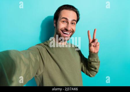 Foto von beeindruckt Bart Millennial Kerl tun Selfie zeigen V-Zeichen tragen Khaki Sweatshirt isoliert auf blauem Hintergrund Stockfoto