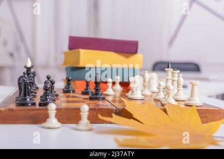 Altes Holzbrett mit Schachfiguren steht auf weißem Tisch. Das Spiel hat begonnen, erste Züge wurden gemacht. In der Nähe liegt trockenes, gelbes Ahornblatt. Seitenansicht. Stockfoto