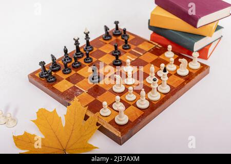 Altes Holzbrett mit Schachfiguren steht auf weißem Tisch. Das Spiel hat begonnen, erste Züge wurden gemacht. In der Nähe liegen Stapel von bunten Büchern. Stockfoto