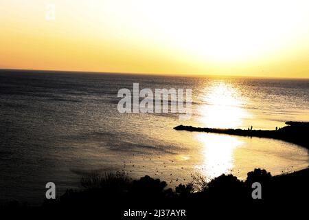 Sonnenuntergang mit Meer in der Bucht, Menschen und Vögel am Strand, Drohnenansicht, Horizont-Linie, Silhouette von Bäumen und Landschaft, kontrastreiche Aufnahme Stockfoto