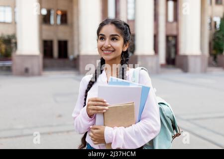 Porträt einer fröhlichen indischen Studentin im Freien mit Rucksack und Arbeitsbüchern, die in der Nähe des College-Gebäudes stehen Stockfoto