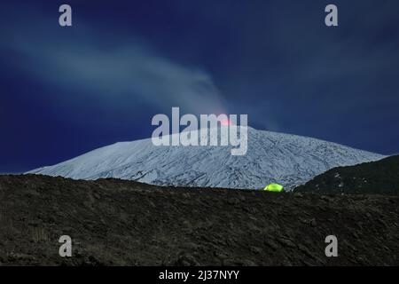 Beleuchtete Zelt am Rande eines alten Krater vor dem schneebedeckten Ätna, die vulkanischen Dampf emittieren, Ätna Park - Sizilien Stockfoto