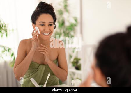 Glückliche Millennial Frau trägt Handtuch, Anwendung Gesichtswasser mit kosmetischen Schwamm in der Nähe des Spiegels zu Hause, leeren Raum Stockfoto