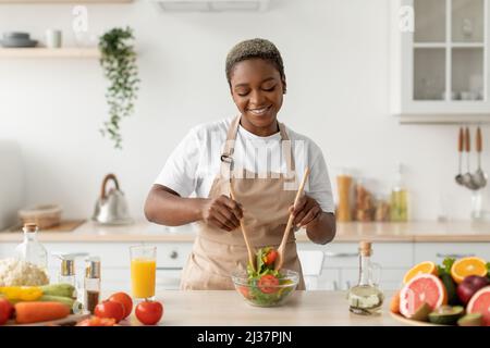 Zufriedene tausendjährige afroamerikanische Dame in Schürze, die Bio-Gemüsesalat am Tisch in der modernen Küche zubereitet Stockfoto