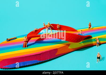 Einige Miniatur-Männer, die Badeanzug tragen, auf einem Regenbogen-gemusterten Flip-Flop vor blauem Hintergrund Stockfoto