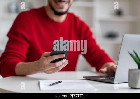 Arabischer Mann Mit Modernem Mobiltelefon, Während Er Am Schreibtisch Mit Laptop Sitzt Stockfoto
