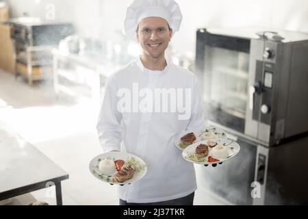 Porträt eines jungen, gutaussehenden, lächelnden Küchenchefs in Uniform, der mit Fertiggerichten in der Küche steht. Fröhlicher kaukasischer Koch, der im Restaurant arbeitet. Konzept der beruflichen Tätigkeit und Haute Cuisine Stockfoto