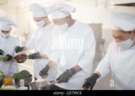 Multirassisches Team von Köchen kocht in weißer Uniform zusammen in der Küche. Asiatische, lateinische und europäische Jungs kochen zusammen. Köche tragen Gesichtsmasken und Schutzhandschuhe Stockfoto