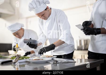 Mehrere Rassen Gruppe von Köchen, die Hauptgerichte beenden, während sie zusammen in der Küche arbeiten. Das Team bereitet Mahlzeiten für das Restaurant zu Stockfoto