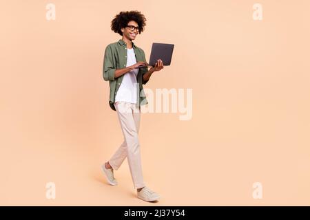 In voller Länge Körpergröße Ansicht der attraktiven fröhlich fokussiert qualifizierten Kerl gehen mit Laptop isoliert über beige Pastellfarben Hintergrund Stockfoto