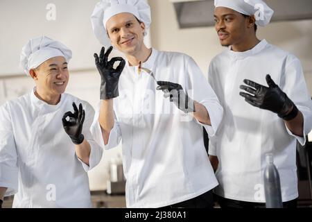 Drei Köche mit unterschiedlichen Nationalitäten verkosten gekochte Gerichte und erfinden neue Gerichte für ein Menü. Kaukasischer Koch mit lateinischen und asiatischen Köchen in Uniform, die zusammenarbeiten. Konzept der Teamarbeit und hohe Küche Stockfoto