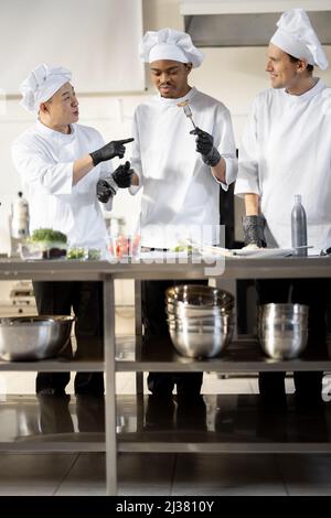 Drei Köche mit unterschiedlichen Nationalitäten verkosten gekochte Gerichte und erfinden neue Gerichte für ein Menü. Asiatischer Koch mit lateinischen und europäischen Köchen in Uniform, die zusammenarbeiten. Konzept der Teamarbeit und hohe Küche Stockfoto
