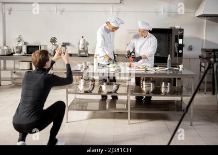 Mann zeichnet am Telefon den Prozess des Kochens von zwei Köchen in einer professionellen Küche auf. Lateinamerikanische und asiatische Köche bereiten Speisen für soziale Medien zu. Konzept des kulinarischen Videobloggens Stockfoto