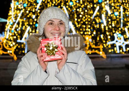 Eine Frau in weißer Winterkleidung steht vor einem Weihnachtsbaum, hält eine rote Geschenkbox und lächelt. Stockfoto