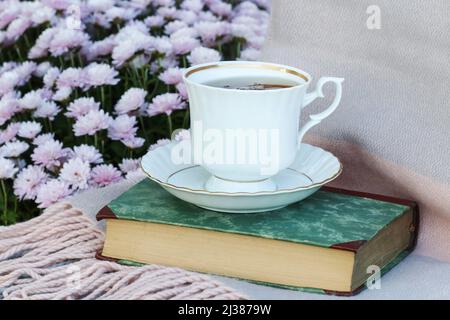 Eine Tasse Tee steht auf einem alten Buch. Im Hintergrund sind rosa Chrysantheme Blüten Stockfoto