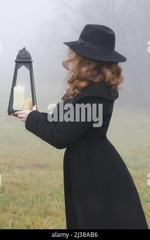 Die rothaarige Frau hält eine eiserne Laterne mit Kerze in ihren Händen. Neblige Landschaft im Hintergrund. Stockfoto